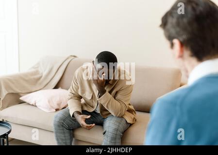 Frustrierter junger afroamerikanischer Mann, der auf dem Sofa sitzt und die Hände in der Nähe des Gesichts im Büro bei einer Sitzung mit einem Psychologen oder Psychiater hält. Verzweifelte BL Stockfoto