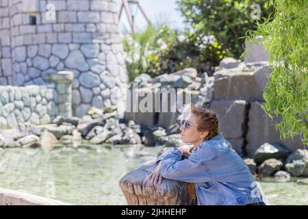 Eine Frau sitzt im Schatten der Bäume und lehnt sich auf ein Holzgeländer vor dem Hintergrund einer Steinburg und Wasser. Friedliche Entspannung im Vergnügungsland Stockfoto