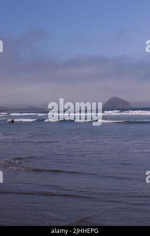 Ein ruhiger, leicht bewölktes Nachmittag an einem Strand in der Nähe von Cayucos, CA mit wenigen Menschen und sanften Wellen. Morro Bay liegt in der Ferne mit seinem riesigen Felsen. Stockfoto