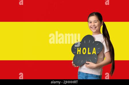 Nettes kleines asiatisches Mädchen mit einer Tafel mit Text HOLA (HALLO) gegen die Flagge Spaniens Stockfoto