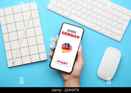 Weibliche Hand hält Handy mit Text DEUTSCH lernen am Bildschirm am Arbeitsplatz, Draufsicht Stockfoto