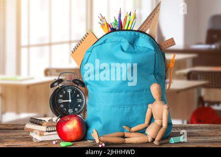 Schulrucksack mit Schreibwaren, menschlicher Holzfigur, Uhr und Apfel auf dem Tisch im Klassenzimmer Stockfoto