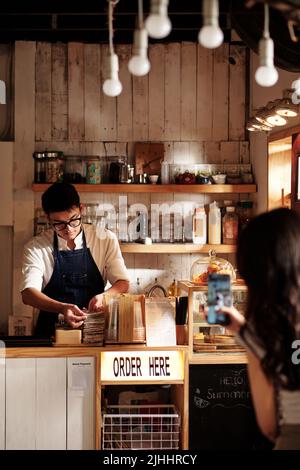 Die verliebte junge Frau filmt heimlich Barista, die am Coffeeshop arbeitet Stockfoto