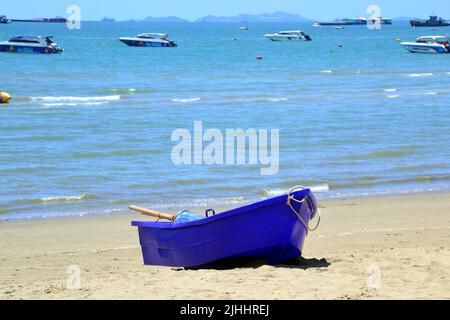 Ein kleines, leeres, blaues Plastikruderboot sitzt am Sandstrand im Badeort Pattaya, Thailand, Asien. Schnellboote im Meer in der Ferne. Stockfoto