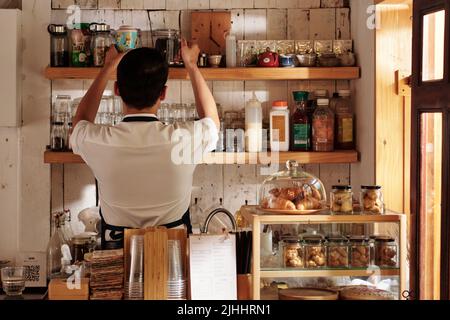 Coffee Shop Barista nimmt Gläser mit schwarzem Tee aus dem Regal, um Getränke für den Gast zuzubereiten Stockfoto