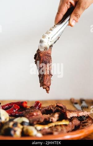 Ein Stück Fleisch in einem tong wird gerade auf einen Teller gelegt. Hochwertige Fotos. Gebratene Zwiebeln, Fleischstücke und Würste in einer Schale aus Ton. Weißer Hintergrund. Hand mit einer Zange Stockfoto