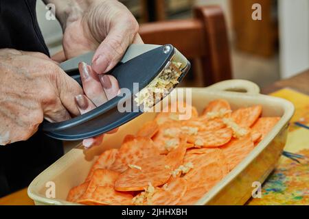 Der Koch drückt den Knoblauch auf Sweet Potato Herb Gratin, bevor er im Ofen gebacken wird. Französische Gourmetküche Stockfoto