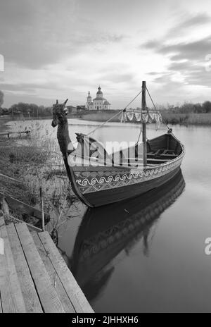 Boot auf dem Fluss Kamenka. Tourist drakkar in der Nähe des Ufers an der Iljinsky Kirche. Susdal, Russland, 2022 Stockfoto