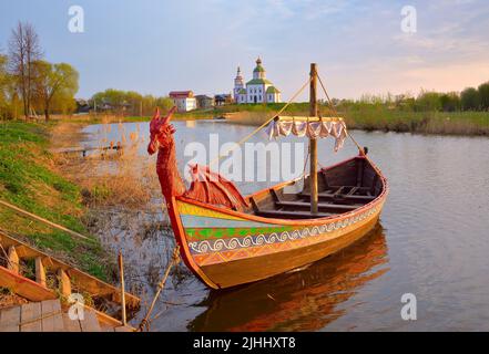 Boot auf dem Fluss Kamenka. Tourist drakkar in der Nähe des Ufers an der Iljinsky Kirche. Susdal, Russland, 2022 Stockfoto