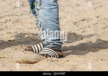 Schwarze Füße in Flip-Flops am Strand mit zerlumpten blauen Jeans Stockfoto