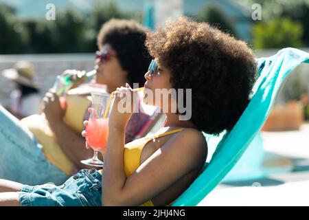 Junge Frauen aus der Bira tragen Sonnenbrillen und trinken Cocktails, während sie auf Liegestühlen auf einer Poolparty liegen Stockfoto