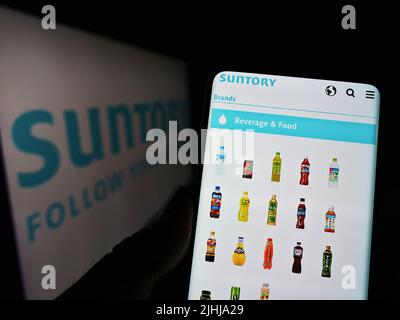 Person, die Mobiltelefon mit Website des japanischen Getränkeunternehmens Suntory Holdings K.K. auf dem Bildschirm mit Logo hält. Konzentrieren Sie sich auf die Mitte des Telefondisplays. Stockfoto