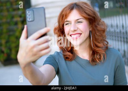 Frau mit roten Haaren lächelt und schaut auf den Bildschirm zu ihrem Handy im Freien Stockfoto