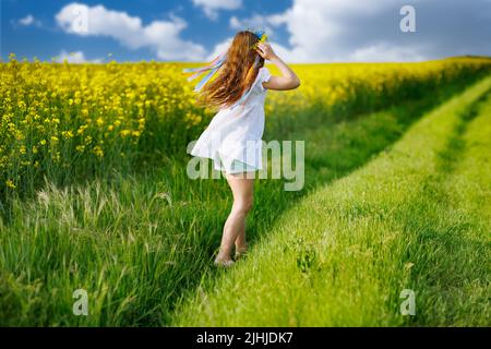 Schönes, energiegeladenes, verträumtes Teenager-Mädchen in einem weißen Kleid und einem leuchtend ukrainischen Kranz, das durch gelb blühende Rapsfelder und grüne Äcker läuft Stockfoto