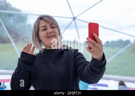 Eine lächelnde asiatische Frau in einer schwarzen Jacke nutzt ein Smartphone für die Online-Videokommunikation und winkt mit der Hand, um Freunde zu grüßen. Online-Kommunikation mit Stockfoto