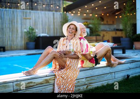 Glückliches Seniorenpaar, das beim Entspannen Getränke genießt und im Sommer am Pool sitzt und die Kamera anschaut. Stockfoto