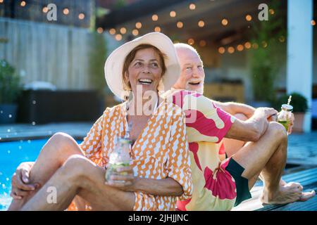 Glückliches Seniorenpaar, das im Sommer beim Entspannen Getränke genießt und am Pool sitzt. Stockfoto