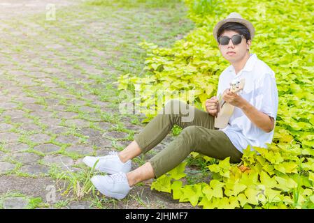 Asiatischer Mann mit Hut, der im Freien Gitarre Ukulele spielt Stockfoto