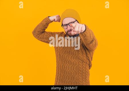 Stilvoller Hipster, der seine Muskeln anbiegend und mit einem Finger auf die Kamera über den orangefarbenen Hintergrund zeigt – körperliches und mentales Kraftkonzept. Hochwertige Fotos Stockfoto