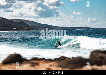 Wilde felsige Küste des Surfspots La Santa Lanzarote, Kanarische Inseln, Spanien. Surfer reiten eine große Welle in felsiger Bucht, Vulkan Berg im Hintergrund. Stockfoto