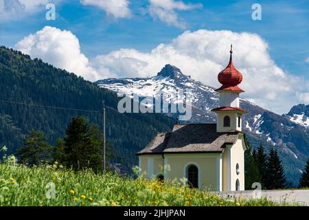 Eine kleine Kapelle auf einer grünen Wiese am Straßenrand einer Landstraße in den Alpen im Sommer Stockfoto