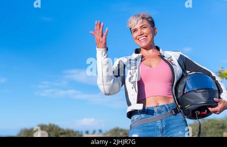 Die junge, hübsche blonde Frau mit kurzen Haaren, die als Biker mit einer Lederjacke und ihrem Schutzhelm gekleidet ist, lacht beim Blick auf die Kamera Stockfoto