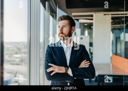 Erfolgreicher Geschäftsmann in formeller Kleidung, der mit gefalteten Armen im modernen Coworking-Büro steht und durch das Fenster schaut Stockfoto