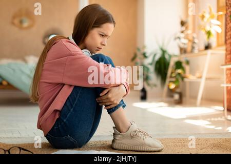 Deprimiertes Teenager-Mädchen, das auf dem Boden sitzt und ihre Knie umarmt, an Cybermobbing leidet und online belästigt wird Stockfoto