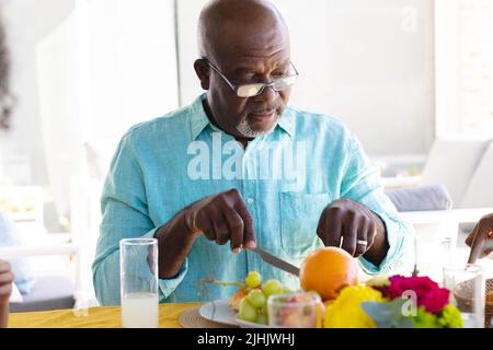 afroamerikanischer kahlköpfiger Mann mit Brille, der zu Hause auf dem Esstisch Früchte isst Stockfoto