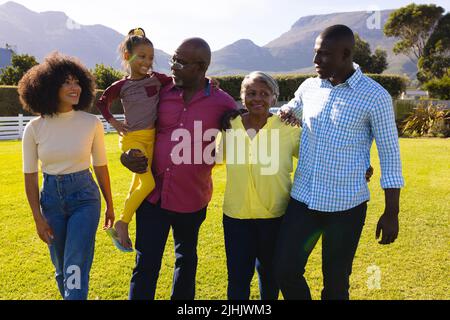 Glückliche multirassische Familie mit mehreren Generationen, die auf einem grasbewachsenen Feld gegen Berge im Hof läuft Stockfoto