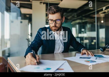 Konzentrierter, gutaussehender Geschäftsmann in einer Brille, der mit Dokumenten im Büroinnenraum arbeitete, Papiere überprüfte und unterschrieb Stockfoto