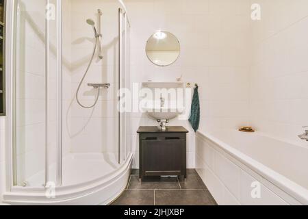 Badewanne mit Glasdusche in der Ecke neben Waschbecken und Spiegel im kleinen hellen Badezimmer mit weißen Kachelwänden Stockfoto