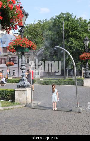 Niedliche kleine Mädchen genießen es, sich von öffentlichen Verneblern auf einem Stadtplatz abzukühlen; Wasserkühlung, Nebelkühlung, Wassersprüher Stockfoto