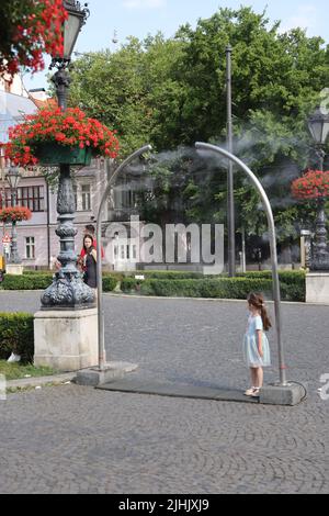 Niedliche kleine Mädchen genießen es, sich von öffentlichen Verneblern auf einem Stadtplatz abzukühlen; Wasserkühlung, Nebelkühlung, Wassersprüher Stockfoto