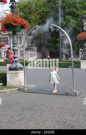 Niedliche kleine Mädchen genießen es, sich von öffentlichen Verneblern auf einem Stadtplatz abzukühlen; Wasserkühlung, Nebelkühlung, Wassersprüher Stockfoto