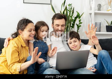 Virtuelles Meeting. Vierköpfige Familie winkt beim Einsatz eines Laptops für Videoverbindungen mit Großeltern oder Familie auf den Bildschirm. Multirassische Mutter, Vater und zwei Kinder winken in der Webcam des Laptops, Videoanruf Stockfoto