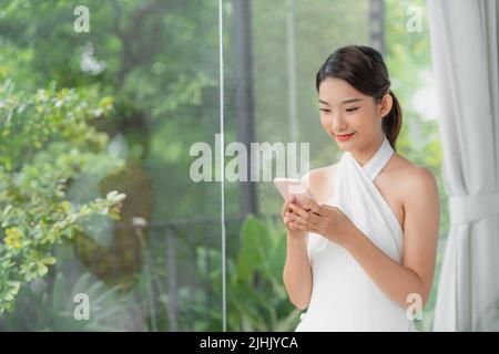 Das asiatische Mädchen sitzt am Tisch neben dem Fenster, hält ein Mobiltelefon und schaut auf den Bildschirm Stockfoto