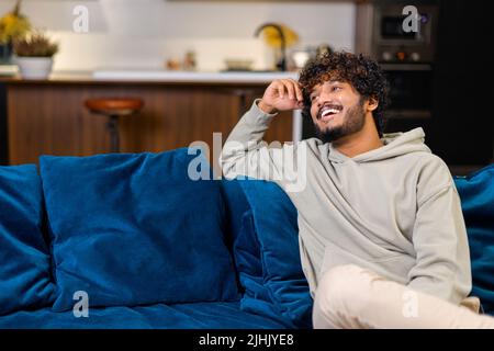 Ethnischer unrasierte Mann, der auf dem blauen gemütlichen Sofa sitzt und lächelt, während er sich wohlfühlt und sich entspannt. Kerl schaut weg. Seine moderne Wohnung im Hintergrund Stockfoto