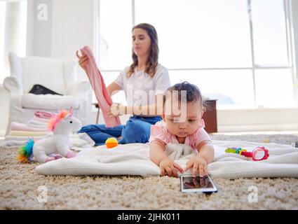 Das wird immer mehr zu einem Kampf jeden Tag. Eine Mutter faltet Kleidung, während ihr Baby in der Lounge spielt. Stockfoto