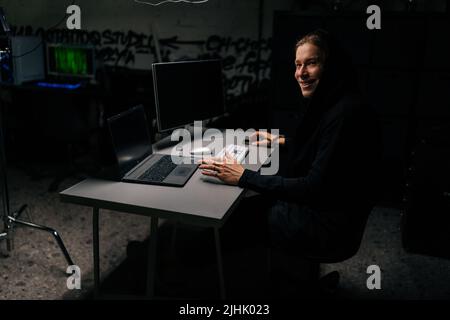 Lächelnder kaukasischer Hacker-Mann in der Haube, der mit einem Laptop und Computern im dunklen Kellerraum am Schreibtisch in die Datenserver des Unternehmens einbrach. Stockfoto