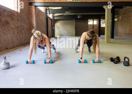 Zwei junge attraktive Frauen tun Liegestütze innen mit Hanteln. Fitness-Frau, die in einem Cross-Fit-Fitnessstudio trainiert. Fitnesskonzept. Hochwertige Fotos Stockfoto