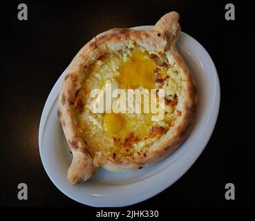 Khachapuri Adjaruli (Georgian Cheese Bread Boat) aus Brot, Käse, Butter und Eigelb auf einem weißen Teller mit schwarzem Hintergrund Stockfoto