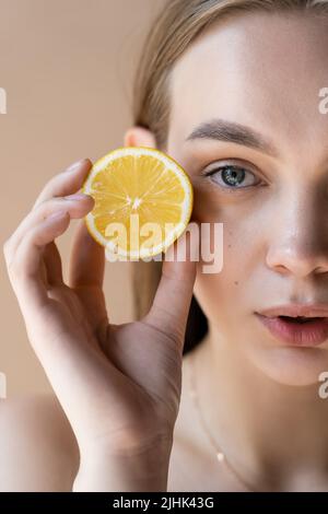 Beschnittene Ansicht der Frau mit natürlichem Make-up, die eine halbe Zitrone isoliert auf Beige hält Stockfoto