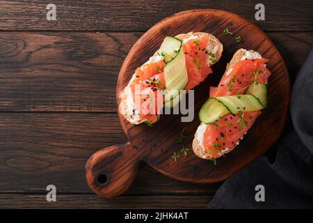 Sandwiches. Lachs-Toast mit Frischkäse, Gurke, schwarzem Sesam und Microgreens auf altem Holztisch-Hintergrund. Meeresfrüchte. Gesunde Ernährung. Fotografie Stockfoto
