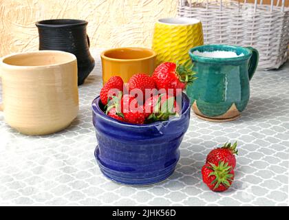 Keramikbecher in verschiedenen Farben und Größen mit Erdbeer-Nahaufnahme. Mehrfarbige Cups mit speziellen glasierten bedeckt. Keramikgeschirr mit glänzender Brandung Stockfoto