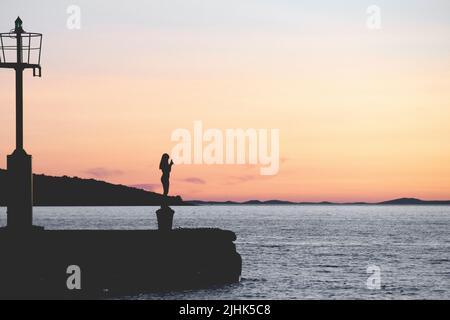 Frau Silhouette steht auf Schiff Anlegestelle in der Nähe des Leuchtturms halten Telefon und fotografieren den schönen Sonnenuntergang der adria Stockfoto