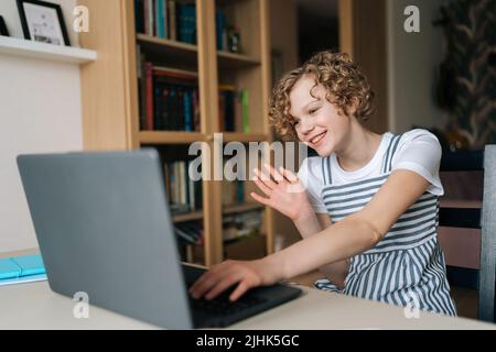 Portrait von fröhlichen Mädchen am Tisch sitzen mit Videoanruf, Blick auf Laptop-Display und winken Hand an Freunde, Familie, Eltern, Lehrer. Stockfoto