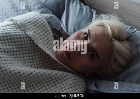 Draufsicht auf depressive blonde Frau mit Menopause, die unter der Decke im Bett liegt Stockfoto