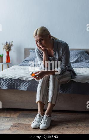 Verärgert blonde Frau mit Wechseljahren hält Flasche mit Pillen und sitzt auf dem Bett Stockfoto