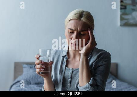 Blonde Frau mit Wechseljahren, die unter Kopfschmerzen leidet und ein Glas Wasser hält Stockfoto